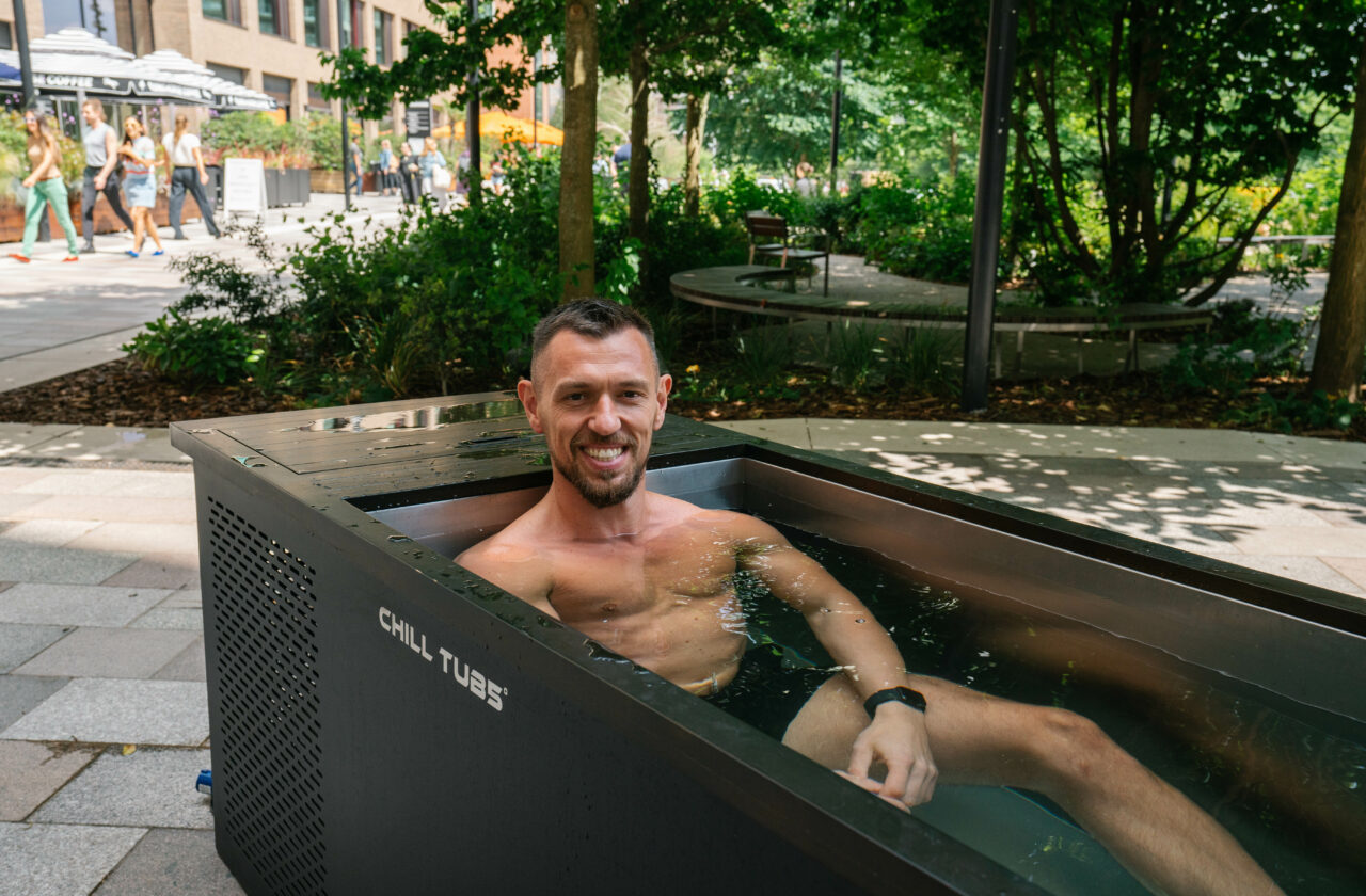Rob Carlin sat in a Chill Tub ice bath on a high street. There are various restaurants in the background of the image, and Rob is smiling directly at the camera.